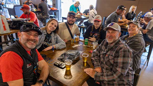 A group of smiling race fans sit around a wooden table inside a casual lounge area, holding drinks and wearing NASCAR apparel and hats. Other guests are seated at tables in the background, creating a lively, social atmosphere.